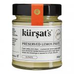 A clear glass jar of Kursat’s preserved lemon paste sits centered on a neutral background. The bright, glossy yellow paste shows flecks of rind and a slightly coarse texture. A blue-and-white label with cursive logo and weight wraps the jar; a metal lid seals it. Soft reflections appear on the glass