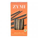 A row of thin golden-brown breadsticks flecked with dark green spinach and tiny dill and onion pieces lies beside a partially opened green-and-white bag labeled Zymi Bakery. Crumbs scatter on a rustic wooden board under soft natural light, highlighting textures. A clear window, shows flecks of herbs.