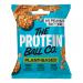 A small resealable brown pouch sits against a white background. The label shows bold brand text and product name, with green and white accents and small icons for vegan and vitamins. A clear window reveals round peanut-brown protein balls, with a few balls scattered beside the bag. Close up details.