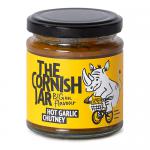 A tight group of six glass jars displays deep-red, chunky hot garlic chutney, visible through clear glass. Each jar wears a cream label with green Cornish script and an illustrated clove of garlic, finished with black metal lids and neat shelf-ready alignment against a plain background. soft studio.