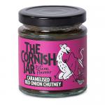 A glass jar of deep mahogany caramelised red onion chutney shows a glossy, chunky texture. It has a pale gold metal lid and a cream label with green logo and elegant script. The jar sits on a neutral surface and catches soft studio reflections. Shallow shadow falls to one side, showing jewel swirls.