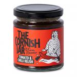 A squat glass jar holds a deep red tomato and chilli relish with visible seeds and glossy tomato pieces. The jar sports a cream label with black and red lettering reading The Cornish Jar, floral accent, and a silver twist-off lid. Light reflects on the glass. Smooth, slightly coarse texture close-up