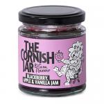 Six glass jars sit in a neat row, each filled with jewel-toned spreads: deep blackberry purple, soft apple gold, and creamy vanilla amber. Labels display The Cornish Jar logo and flavor names, while metal lids catch light, and textured fruit imagery decorates each label. on a pale wooden surface lit
