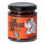 A clear glass jar holds deep red, glossy chilli jam with visible chili flecks. A cream label wraps the jar, featuring a black-and-white Cornish lighthouse logo, bold serif brand name and smaller product text. A silver metal lid seals the jar. The jam shows tiny seeds and a glossy, translucent sheen.