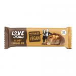 A milk-chocolate–coated peanut caramel bar sits beside its wrapper labeled LoveRaw. The wrapper uses earthy browns and cream, with peanut and caramel graphics. A clean cross-section shows glossy caramel and crunchy peanuts atop a dense nougat, and sits on a white background.
