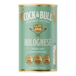 The photo shows a glass jar of red bolognese sauce with visible meat pieces. The label features a rooster and bull illustration and bold typography. A metal lid gleams, the jar sits on a wooden surface under soft studio lighting with shallow depth of field. Soft condensation dots the interior glass.