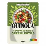 A clear packet of Quinola green lentils fills the frame, its transparent window revealing small pale green legumes. A cream-and-green label shows the brand name and product title; loose lentils scatter across a light wooden surface beside the packet. Soft natural light casts soft shadows and bokeh.