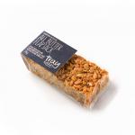 Photo shows a stack of rectangular golden flapjacks, each with visible rolled oats and a glossy butter glaze. One bar sits unwrapped on a wooden board, revealing a dense, chewy oat texture and slightly crumbed edges; a box of twelve bars sits in the background.