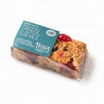 A single Paradise flapjack sits on a tray amid eleven identical bars, each rectangular and golden-brown with visible oats and a glossy syrup glaze. Clear wrapper displays bright bakery branding and a product label, while the tray forms tidy rows under soft lighting.