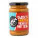 Photo shows six glass jars of Yumello Crunchy Smoky Harissa Peanut Butter, each 285 g, lined in two rows. Labels display bold orange-red harissa graphics and brand name; lids are matte black. Slightly grainy peanut butter peeks through the clear glass, casting warm reflections. Soft lighting detail.