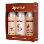 Three compact 200ml glass bottles sit in a recyclable cardboard tray, each labeled with colorful minimalist branding. Clear viscous syrup shows rich amber, ruby and golden hues. Metal screw caps and neat nutritional panels create a tidy artisanal grocery aesthetic. Soft lighting reveals label sheen.
