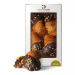 A close-up photograph shows four round Rocher chocolates arranged in a neat row inside a partially open Di Santo & Family Bakery box. Each confection displays a glossy chocolate coating studded with chopped hazelnut pieces, set against a simple cream insert and rustic branded packaging. in soft glow