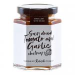 A neat row of six glass jars sits on a pale surface. Each jar shows chunky, deep red sun-dried tomato and garlic chutney. Cream labels with black text and a small logo circle the jars; metal lids reflect soft studio lighting.
