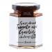 A neat row of six glass jars sits on a pale surface. Each jar shows chunky, deep red sun-dried tomato and garlic chutney. Cream labels with black text and a small logo circle the jars; metal lids reflect soft studio lighting.