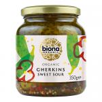 Clear glass jars labeled Biona Organic Gherkins show plump, bright-green gherkins packed in pale brine. Simple white labels with green accents and organic icons encircle the jars. The arrangement sits on a neutral surface against a soft, light background.