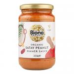 A glass jar with a white screw-top lid displays deep reddish-brown satay sauce. A green-and-orange label shows the Biona logo and product name, with an illustration of peanuts and a red chili. The label includes organic branding and weight text. Glass shows glossy reflections and fine print labels.