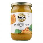 A tall glass jar with a warm orange label shows bold white lettering and a small pumpkin illustration. The jar reveals thick, vibrant pumpkin soup with visible spice flecks. A wooden spoon beside the jar scoops smooth, dense puree. Soft light catches the glossy surface and scattered toasted seeds...