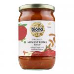 A clear glass jar shows deep red minestrone with visible beans, diced carrots, pasta and green herbs. A green-and-white Biona label wraps the jar, displaying organic branding and product name. The jar sits on a light wooden surface against a neutral backdrop. A silver lid catches gentle studio light.
