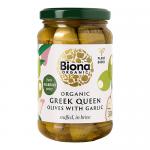 A clear glass jar reveals large green Greek queen olives tightly packed in pale brine, each cavity stuffed with a whole garlic clove. A simple organic label with green and white accents wraps the jar, while condensation and reflective highlights give a fresh, glossy appearance. They catch the light.