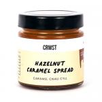 A glossy glass jar with a screw lid displays swirled hazelnut-caramel spread. A bold label wraps the jar with warm brown and amber tones, brand name centered. Toast crumbs and scattered hazelnuts border the jar on a neutral background. Soft studio light reveals glossy swirls and warm color gradient.