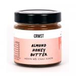 A neat row of six glass jars of Crwst Almond Honey Butter (210g) shows golden, creamy spread visible through clear glass, dotted with tiny almond flecks. White minimalist labels read CRWST Breakfast Spreads; gold lids reflect soft natural light on a pale surface Subtle shadows add depth crisp edges