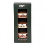 A neat gift set displays six small glass jars in two rows inside a kraft box, each jar showing a colorful label and creamy spread visible through the glass. The box lid rests beside the tray, a linen ribbon and shredded paper filling add rustic texture to the scene. Soft light highlights lids gloss.