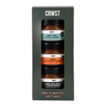 Three small glass jars of glossy caramel sit nestled in a kraft gift box filled with crinkled paper. Each jar features a clean CRWST label and metallic lid; warm light catches amber swirls and a velvety, slightly sticky texture against a neutral background. Soft shadows add depth and artisanal charm