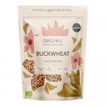 A simple kraft paper pouch with a clear window shows light-brown triangular buckwheat kernels piled beside a small cluster of mixed dried ingredients—orange carrot flakes and green herb bits—on a plain white background, soft natural lighting highlighting textures. Subtle shadows create gentle depth.