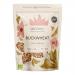 A simple kraft paper pouch with a clear window shows light-brown triangular buckwheat kernels piled beside a small cluster of mixed dried ingredients—orange carrot flakes and green herb bits—on a plain white background, soft natural lighting highlighting textures. Subtle shadows create gentle depth.