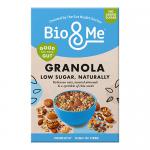 Front-facing Bio&Me granola pack rests on a pale wooden surface under soft daylight. Cream and sage-green packaging features a clear window revealing golden oat clusters studded with seeds and dried berries. A white ceramic bowl and silver spoon display crunchy clusters; loose oats scatter nearby..