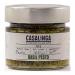 A clear glass jar reveals bright green basil pesto with a slightly coarse, oily texture. A cream-and-red Casalinga label wraps the jar, showing the product name and Italian motifs. A metal lid tops the jar; a wooden spoon and loose basil leaves sit beside it on a neutral surface. Soft light accents.