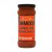 Glass jar with a bright red-orange curry sauce fills the frame; flecks of mustard seed and chilli flakes dot the viscous surface. A colorful label shows the brand name and product details, while a silver lid catches light and a small spoon rests beside the jar. Condensation beads cling to the glass;