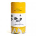 A matte gift tin labeled 'Good Morning Sunshine' and 'Dark Woods' sits on a weathered wooden table. Warm brown and gold label tones contrast with scattered coffee beans and a small spoon. Soft morning light casts long gentle shadows across the scene. Matte tin shows a small embossed logo and weight.