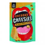 A torn yellow-orange packet labeled Cheesies Cheddar Crunchy Popped Cheese sits on a wooden board. Nine irregular, airy cheddar-orange puffs spill from the bag, their textured surfaces dusted with fine cheese powder. Crumbs and a blurred charcuterie backdrop frame the scene. Warm glow accents subtly