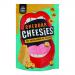 A torn yellow-orange packet labeled Cheesies Cheddar Crunchy Popped Cheese sits on a wooden board. Nine irregular, airy cheddar-orange puffs spill from the bag, their textured surfaces dusted with fine cheese powder. Crumbs and a blurred charcuterie backdrop frame the scene. Warm glow accents subtly