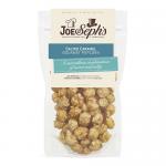 A glossy pouch of Joe & Seph’s Salted Caramel Popcorn sits upright, showing warm golden caramel kernels through a clear window. The brand logo and whimsical illustrations frame the bag; scattered popped corn and salt crystals outline the shiny, sticky coating. Muted kitchen tiles form a soft backdrop