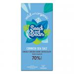 A matte black and cream wrapper frames a 75g extra-dark chocolate bar; a broken square reveals deep, glossy dark brown chocolate studded with mixed seeds and glistening flakes of sea salt. Crisp typography and a coastal motif appear on the packaging. A torn corner shows silver foil & embossed cocoa.