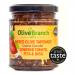 A glass jar of sundried tomato, feta and Greek basil tapenade sits on a weathered wooden board. Deep red spread shows flecks of white cheese and bright green basil, glossy olive oil pooling on top. A sprig of basil and scattered olives garnish the scene sunlit reflections highlight rustic glass lid