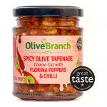 A clear glass jar reveals a coarse, deep red-orange tapenade flecked with chopped Florina peppers, olives, and chilli. A glossy oil sheen catches light. The jar bears a simple Olive Branch label and metal lid, set against a soft neutral background. Warm light falls across a wooden board, and flakes.
