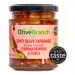 A clear glass jar reveals a coarse, deep red-orange tapenade flecked with chopped Florina peppers, olives, and chilli. A glossy oil sheen catches light. The jar bears a simple Olive Branch label and metal lid, set against a soft neutral background. Warm light falls across a wooden board, and flakes.