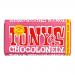A brightly colored wrapper shows bold yellow and red blocks with an oversized handwritten logo. A milk chocolate bar with irregular, chunky segments sits partly unwrapped, revealing caramel and crunchy biscuit pieces. A Fairtrade emblem appears on the lower corner. The metal glints at the torn edges.