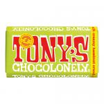 Bright, colorful wrapper displays a bold red Tony's Chocolonely logo over a milk-brown field; creamy hazelnut illustrations and scattered crunchy nibs pattern the packaging while a round Fairtrade badge gleams. The 180g bar lies slightly angled, casting a soft, natural shadow. Light foil sheen fine.