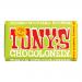 Bright, colorful wrapper displays a bold red Tony's Chocolonely logo over a milk-brown field; creamy hazelnut illustrations and scattered crunchy nibs pattern the packaging while a round Fairtrade badge gleams. The 180g bar lies slightly angled, casting a soft, natural shadow. Light foil sheen fine.