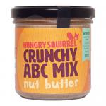 Six identical clear jars with white lids sit in two neat rows. Bright yellow labels show a playful squirrel logo and bold text. Each jar reveals a thick, caramel-hued spread studded with crunchy nut and cereal pieces; a spoon lifts a textured scoop against a clean white backdrop. Soft reflections.