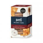 Close-up of a Peter's Yard Spelt & Fig mini cracker pack showing a rustic, pale-brown flatbread studded with dark fig pieces and scattered seeds. Crisp edges and textured surface contrast with simple kraft-style packaging featuring minimalist branding and an illustrated fig motif. Tiny crumbs nearby