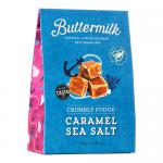 A glossy rectangular sharing box sits open to reveal six neatly stacked 150g caramel-and-sea-salt buttermilk confectionery packs wrapped in cream and bronze foil. The box bears subtle seaside motifs; salted caramel pieces and buttermilk swirls feature in the printed imagery. Soft metallic highlights