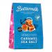 A glossy rectangular sharing box sits open to reveal six neatly stacked 150g caramel-and-sea-salt buttermilk confectionery packs wrapped in cream and bronze foil. The box bears subtle seaside motifs; salted caramel pieces and buttermilk swirls feature in the printed imagery. Soft metallic highlights