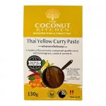The image shows a small glass jar of The Coconut Kitchen Easy Yellow Curry Paste on a neutral surface. Bright yellow paste is visible through the glass. The label is pale yellow with a coconut graphic, ingredient text and a silver metal lid catching soft light. A slight shadow falls beneath the jar.