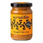Six jars of Butter Bike Co Rugged Peanut Butter (285g) sit on a wooden board. Labels display a bold logo and rustic typography. Matte black lids cap the jars. One jar is open, revealing thick, textured peanut butter with a spoon inside; scattered peanuts and crumbs surround them.