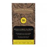 Close-up of a chocolate bar and its wrapper on a rustic wooden surface. The milk chocolate shows glossy, segmented squares and delicate golden flecks suggesting wild gorse flower. A cream-and-gold wrapper with botanical illustrations sits beside scattered yellow gorse blooms. Soft shadows linger on.